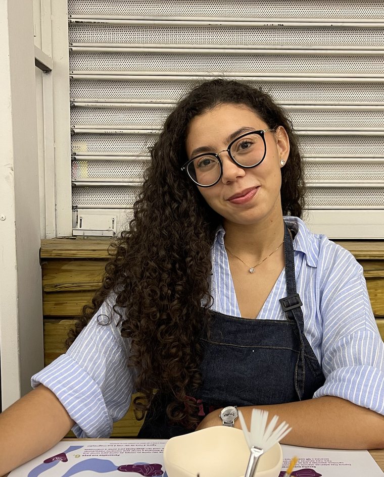 Foto da Eduarda sorrindo, sentada à mesa num ateliê de pintura em cerâmica